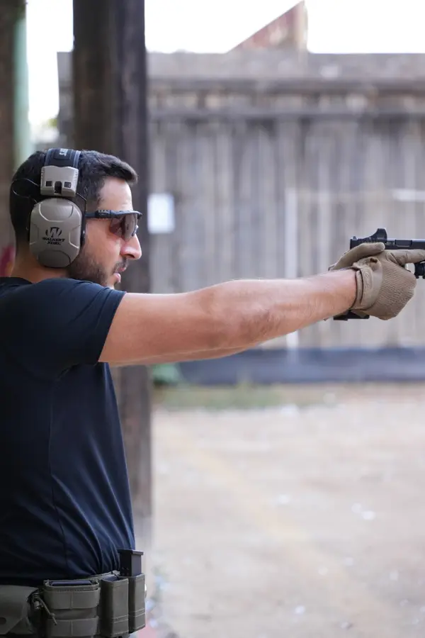 A shooter aiming a pistol at an outdoor range, wearing ear protection, eye protection and shooting gloves