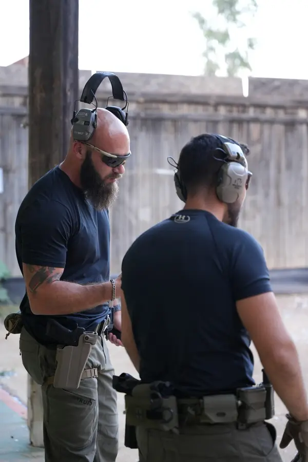 An instructor coaching a trainee on the firing line at an outdoor range, both wearing ear protection